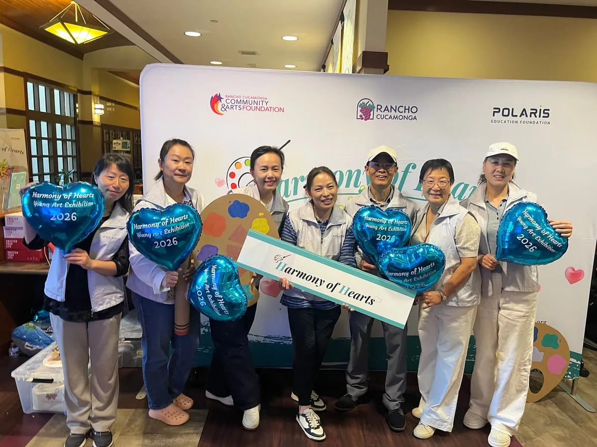 Volunteers in white vests hold blue heart balloons in front of the Harmony of Hearts backdrop.