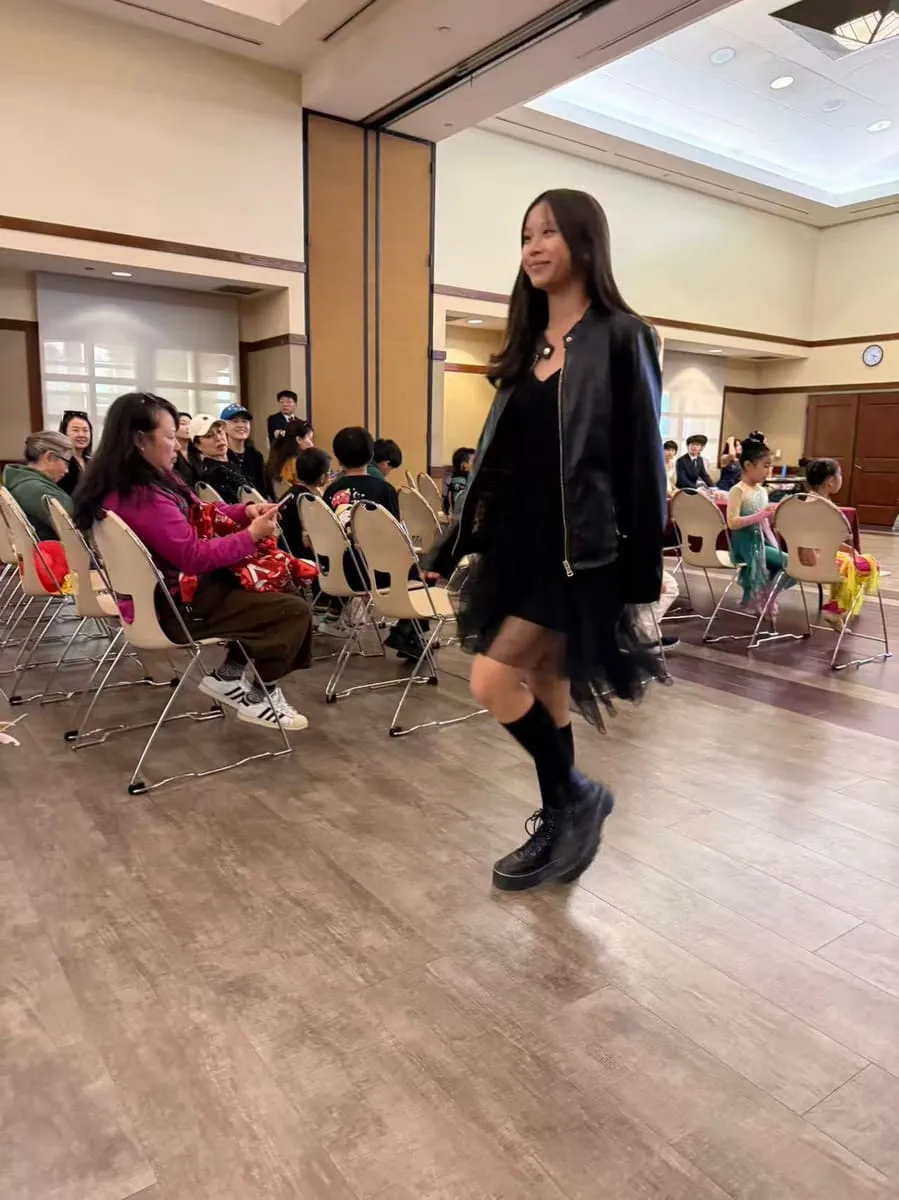 A girl in black walks the runway past rows of seated families.
