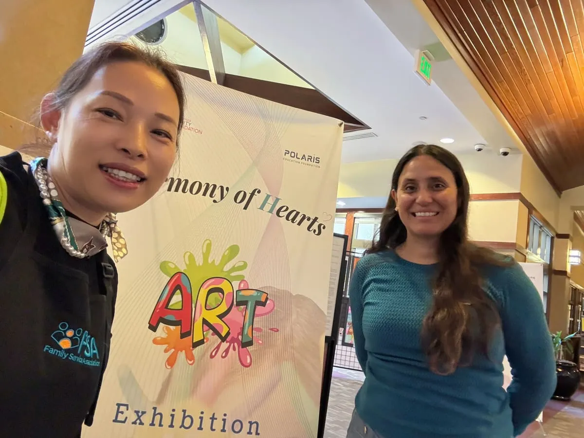 Two women take a selfie beside the Harmony of Hearts Art Exhibition banner.