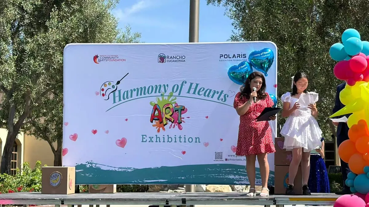 A speaker reads from the stage while a girl in a white dress waits beside her.