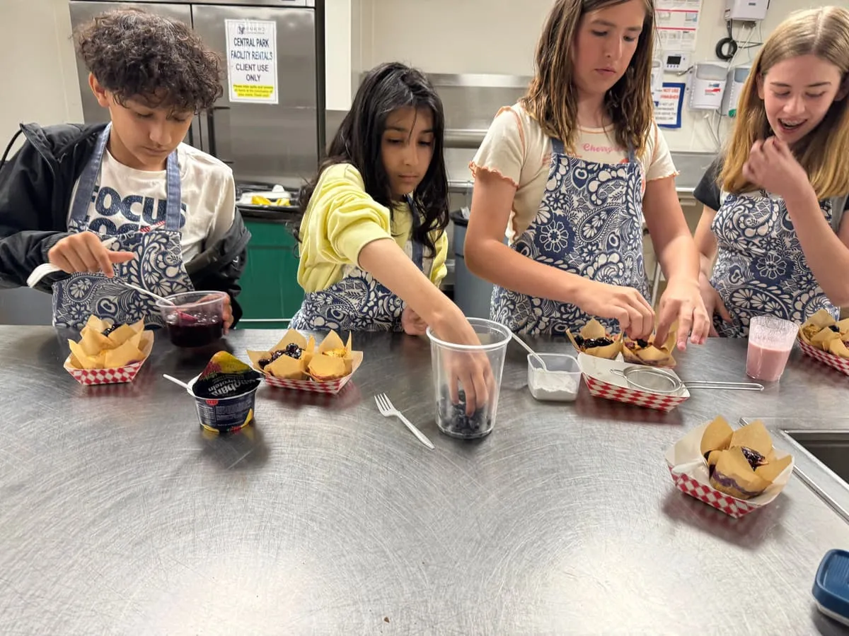 Children assemble snacks at a steel kitchen counter.