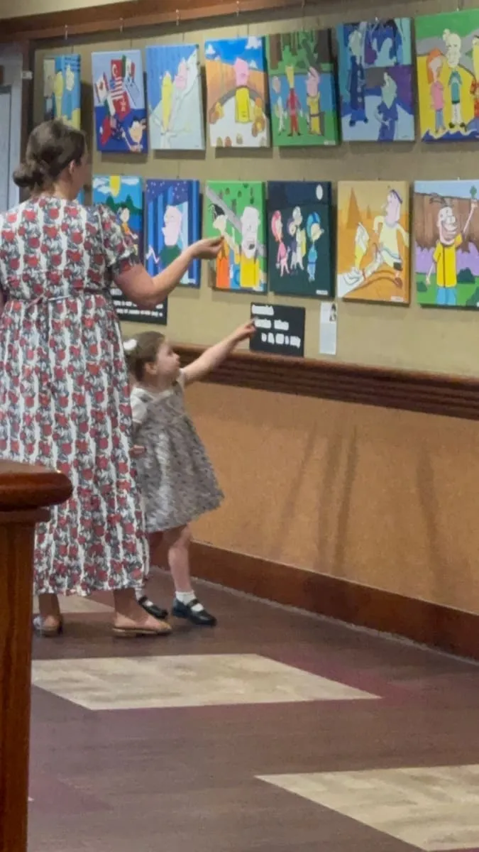 A mother and her small daughter point at paintings on the wall.