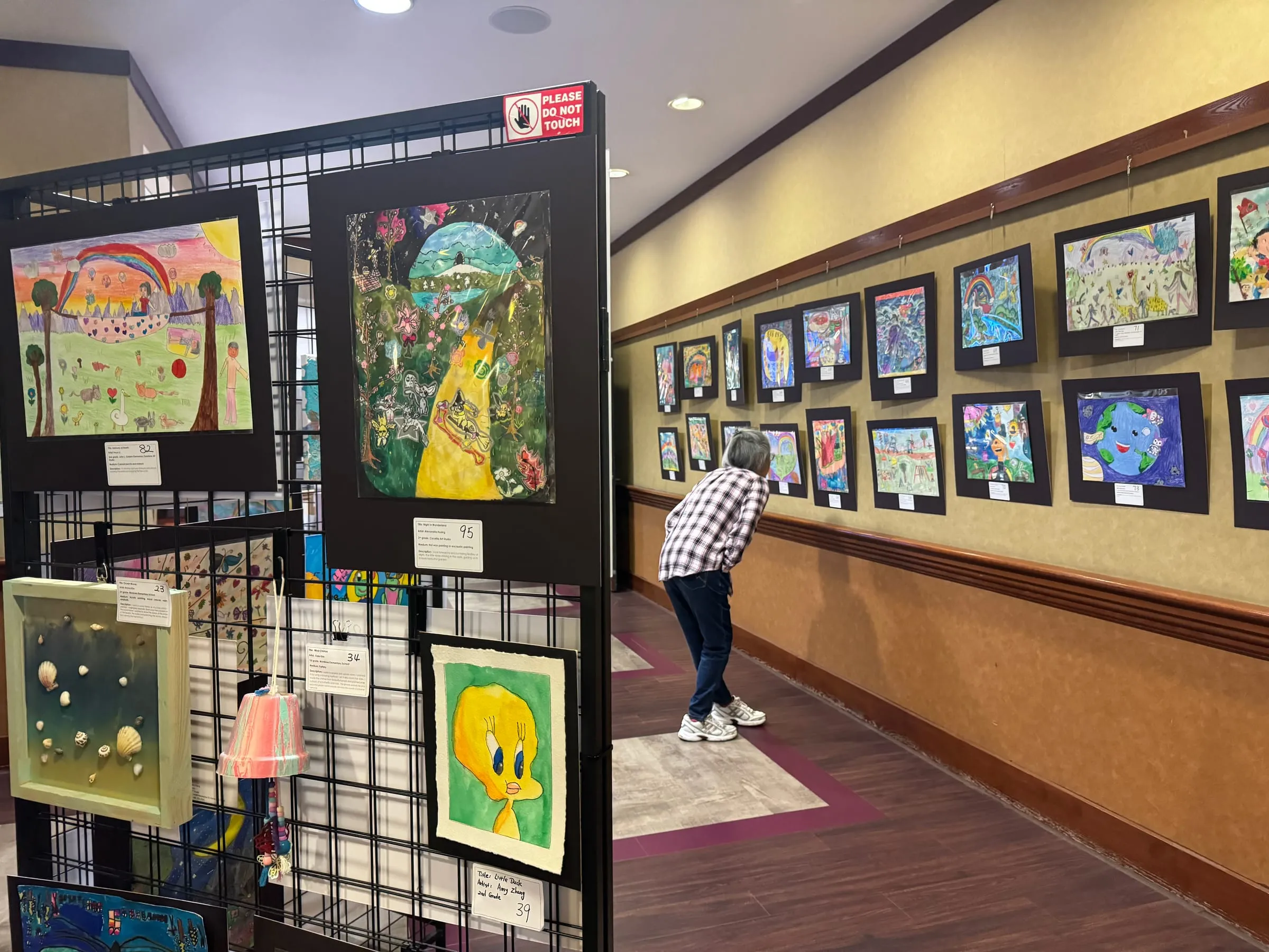 Display panels and a long wall of framed student paintings at the exhibition, with a visitor looking closely at one.