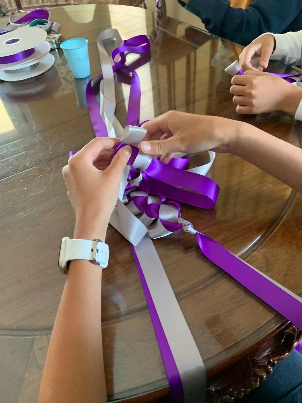Hands braiding purple and silver ribbon into a lei at a table.